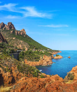 Le Massif de l'Estérel près de Cannes, avec ses roches rouges spectaculaires plongeant dans les eaux bleues de la Méditerranée, et une végétation méditerranéenne luxuriante sous un ciel clair.