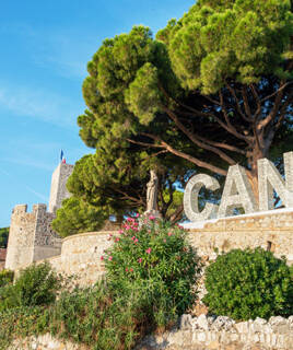 Le Château de la Castre et l'église Notre-Dame d'Espérance à Cannes, avec le célèbre signe "CANNES" en lettres géantes, entourés de végétation méditerranéenne et de pins, sous un ciel bleu clair.