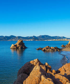 La côte rocheuse près de Cannes, avec des rochers rouges émergeant des eaux calmes de la Méditerranée et une plage de sable en arrière-plan, sous un ciel bleu clair.
