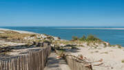 Chemin sablonneux bordé d'une clôture en bois sur les dunes du Banc d'Arguin au Cap Ferret, avec l'océan Atlantique et des bancs de sable au loin sous un ciel bleu clair.