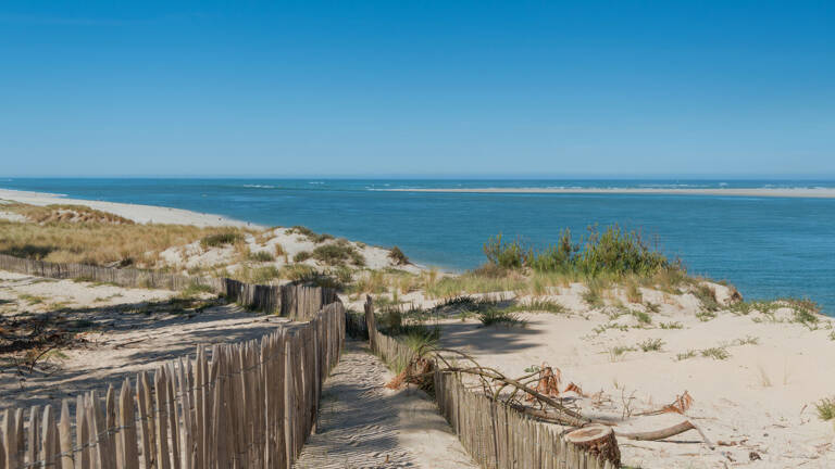 Chemin sablonneux bordé d'une clôture en bois sur les dunes du Banc d'Arguin au Cap Ferret, avec l'océan Atlantique et des bancs de sable au loin sous un ciel bleu clair.