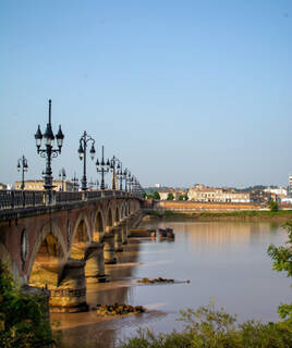 Le Pont de Pierre enjambant la Garonne à Bordeaux, avec ses arches et lampadaires distinctifs, sous un ciel clair et des bâtiments en arrière-plan.