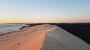 Vue aérienne de la Dune du Pilat au Cap Ferret au coucher du soleil, montrant sa crête s'étendant entre l'océan Atlantique et une forêt dense, avec de petites silhouettes humaines en bas.