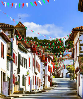 Une rue de village basque décorée de guirlandes aux couleurs rouge, verte et blanche, probablement à Capbreton, avec des maisons traditionnelles blanches aux volets colorés sous un ciel bleu.