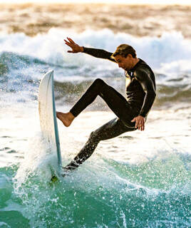 Un surfeur en combinaison sombre chevauchant une vague sur la plage de Capbreton, avec la lumière du soleil en arrière-plan, projetant de l'écume autour de sa planche.