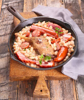 Un plat de cassoulet traditionnel de Carcassonne, servi dans une cocotte en fonte, contenant des haricots blancs, des saucisses, du confit de canard et de la poitrine fumée, posé sur une planche en bois rustique.