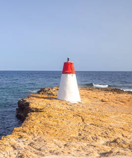 Une petite balise rouge et blanche se dresse sur une formation rocheuse côtière à Carry-le-Rouet, marquant le littoral méditerranéen avec l'océan en arrière-plan sous un ciel bleu clair.