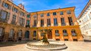 La Place d'Albertas à Aix-en-Provence, avec sa fontaine centrale et ses bâtiments historiques aux façades ocres et aux volets verts, sous un ciel bleu clair.