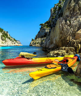 Deux kayaks, un rouge et un jaune, posés sur la plage de galets aux eaux cristallines d'une calanque de Cassis, entourée de falaises rocheuses et de végétation méditerranéenne sous un ciel bleu intense.