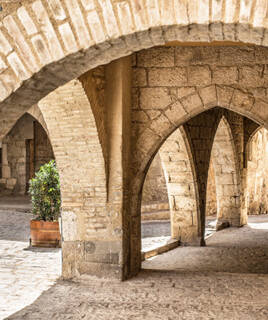 Des arches en pierre du Château de Peralada sur la Costa Brava, s'ouvrant sur une cour pavée avec un pot de fleurs vertes, sous un éclairage naturel.