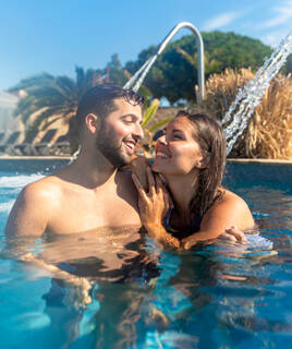 Un couple souriant se détend dans une piscine d'un camping près d'Aix-en-Provence, profitant des jets d'eau sous un ciel bleu ensoleillé.