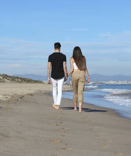 Un couple marchant main dans la main sur une plage de sable près d'Aix-en-Provence, au bord de l'eau, avec des dunes et une ville lointaine en arrière-plan sous un ciel bleu clair.