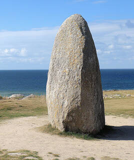 Un imposant menhir solitaire se dresse sur un terrain herbeux au Croisic, avec l'océan en arrière-plan sous un ciel bleu parsemé de nuages blancs.