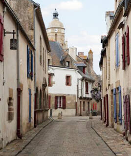 Une ruelle pavée du Croisic, en Loire-Atlantique, bordée de maisons traditionnelles aux façades claires et aux volets colorés, avec un clocher d'église visible au loin sous un ciel nuageux.