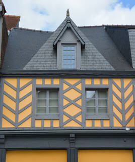 Une façade de maison médiévale à pans de bois, typique de la cité médiévale de Dol-de-Bretagne, avec des motifs géométriques jaunes et des fenêtres à petits carreaux, sous un toit d'ardoise et un ciel nuageux.