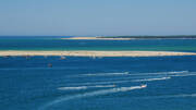 Vue aérienne du Banc d'Arguin depuis la Dune du Pilat, montrant l'île de sable blanc au milieu des eaux bleues du Bassin d'Arcachon, parsemée de bateaux et de sillages.