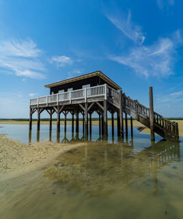 Une cabane tchanquée typique du Bassin d'Arcachon, perchée sur des pilotis dans l'eau peu profonde et le sable, sous un ciel bleu avec quelques nuages.