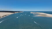 Vue aérienne du Banc d'Arguin en Gironde, un vaste banc de sable au large de la côte, avec des bateaux laissant des sillages sur l'eau bleue et la Dune du Pilat en arrière-plan.