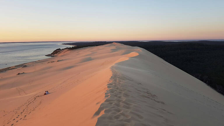 Vue aérienne de la Dune du Pilat en Gironde au coucher du soleil, montrant sa crête s'étendant entre l'océan Atlantique et une forêt dense, avec de petites silhouettes humaines en bas.
