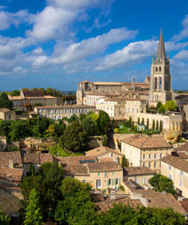 Vue plongeante sur le village médiéval de Saint-Émilion en Gironde, avec son église monolithe et ses toits ocres sous un ciel bleu parsemé de nuages blancs.