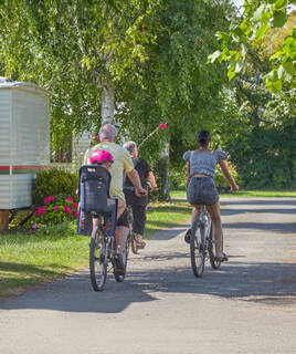 Une famille, dont un grand-parent avec un enfant sur un siège vélo à l'arrière, fait une balade à vélo sur une allée de camping bordée de mobil-homes et d'arbres, près d'Aix-en-Provence.