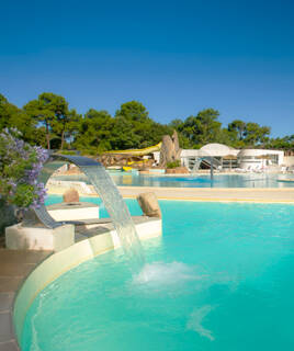 Une piscine de camping spacieuse au Domaine La Yole, avec des cascades d'eau et des toboggans en arrière-plan, entourée de pins et de chaises longues sous un ciel bleu éclatant.