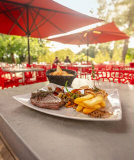 Un plat de viande, frites et légumes garni de romarin, servi sur une table extérieure d'un restaurant, avec des parasols rouges et d'autres tables en arrière-plan, probablement dans un camping près d'Aix-en-Provence.