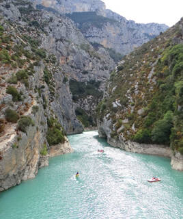Les eaux turquoise des Gorges du Verdon, près de Gréoux-les-Bains, avec des kayaks et des pédalos naviguant entre les falaises escarpées et la végétation méditerranéenne sous un ciel lumineux.