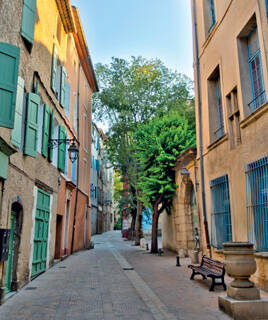 Une rue étroite et pittoresque du village de Gréoux-les-Bains, bordée de bâtiments aux façades colorées et aux volets verts ou bleus, avec un arbre central et un banc en pierre, sous un ciel clair.