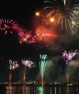 Un feu d'artifice éclatant au-dessus d'un plan d'eau à Grimaud, avec des explosions colorées se reflétant faiblement à la surface, et une ville lointaine illuminée à l'horizon.