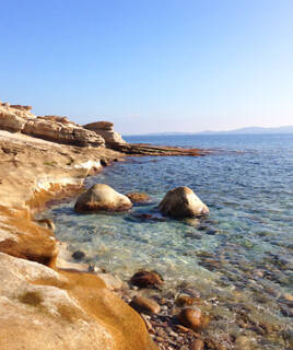 Une crique rocheuse sur la côte varoise près de Grimaud, avec des eaux claires et turquoise baignant des rochers aux couleurs chaudes, sous un ciel bleu dégagé.