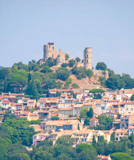 Le village médiéval de Grimaud, avec les ruines de son château et ses tours dominantes, s'étageant à flanc de colline parmi les arbres et les maisons aux toits de tuiles, sous un ciel bleu clair.