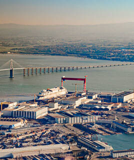 Une vue aérienne des Chantiers de l'Atlantique à Saint-Nazaire, près de Guérande, avec d'immenses navires en construction dans le port, dominés par de grandes grues rouges, et le Pont de Saint-Nazaire en arrière-plan, enjambant l'estuaire de la Loire.