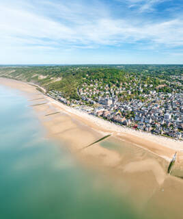 Une vue aérienne de la plage de Veules-les-Roses, près du Havre, avec sa longue étendue de sable, ses brise-lames et la ville côtière nichée entre la mer et les collines verdoyantes, sous un ciel bleu parsemé de nuages.