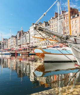 Le vieux port de Honfleur, près du Havre, avec ses bateaux traditionnels amarrés et les reflets des maisons colorées bordant les quais dans l'eau, sous un ciel bleu clair.
