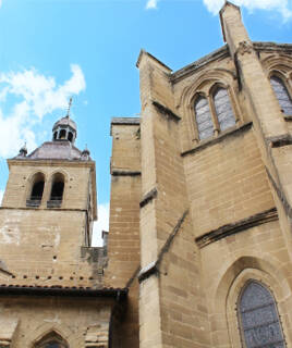 L'Abbaye de Saint-Antoine-l'Abbaye en Isère, une imposante structure médiévale en pierre avec des murs hauts, des fenêtres gothiques et un clocher octogonal, vue d'en bas sous un ciel bleu parsemé de nuages.
