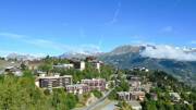 Une vue panoramique sur la station de l'Alpe d'Huez en Isère, avec ses bâtiments nichés dans la montagne verdoyante, et les sommets enneigés des Alpes en arrière-plan sous un ciel bleu clair.