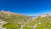 Le Col du Galibier en Isère, un col de montagne avec des routes sinueuses traversant des paysages alpins verdoyants et des sommets rocheux, sous un ciel bleu profond.