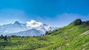 Un paysage montagneux spectaculaire aux Deux Alpes en Isère, avec des pentes verdoyantes au premier plan et des sommets enneigés et nuageux en arrière-plan sous un ciel bleu éclatant.
