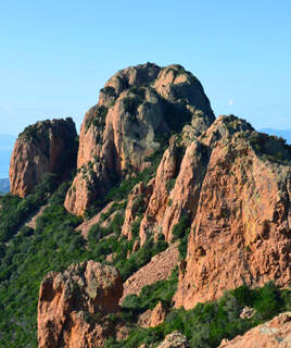 Le Cap Roux dans le Massif de l'Estérel, un promontoire rocheux aux teintes rouges distinctives, couvert de végétation méditerranéenne luxuriante, s'élevant au-dessus de la mer sous un ciel bleu clair.