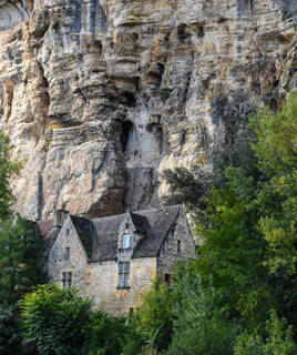 Une maison ancienne en pierre avec un toit en ardoise, nichée au pied de l'impressionnante falaise du Fort Troglodyte de La Roque-Gageac, entourée de végétation.