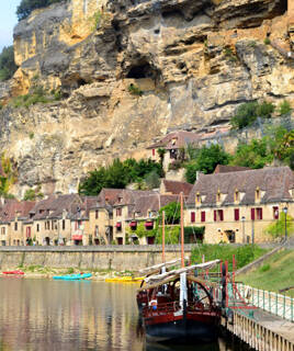 Une gabarre traditionnelle amarrée sur la rivière Dordogne à La Roque-Gageac, avec les maisons du village troglodytique adossées à la falaise en arrière-plan sous un ciel clair.