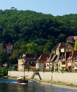 Le village de La Roque-Gageac, en Dordogne, avec ses maisons de pierre aux toits de tuiles rouges le long de la rivière, un bateau naviguant sur l'eau et une colline boisée en arrière-plan sous un ciel bleu clair.