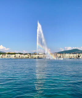 Le Jet d'Eau de Genève, sur le Lac Léman, s'élève majestueusement dans le ciel avec un arc-en-ciel visible, la ville de Genève en arrière-plan et des montagnes lointaines sous un ciel bleu éclatant.