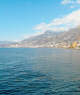 Les rives du Lac Léman, près de Montreux, avec les montagnes en arrière-plan parsemées de neige et de bâtiments, sous un ciel bleu avec quelques nuages.