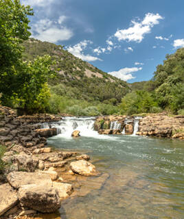 Le fleuve Lez à Lattes, près de Montpellier, avec des rapides et des rochers au premier plan, bordé par des collines verdoyantes sous un ciel bleu parsemé de nuages blancs.