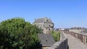 Les remparts de la cité médiévale de Guérande, en Loire-Atlantique, avec une tour de pierre et des toits d'ardoise visibles derrière les arbres, sous un ciel bleu clair.