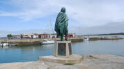 La statue de Pierre Bouguer au Croisic, en Loire-Atlantique, se dresse au bord d'un port avec des bateaux amarrés et des bâtiments en arrière-plan, sous un ciel partiellement nuageux.