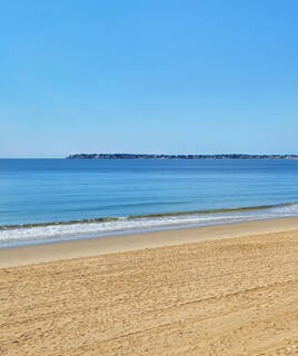 La longue plage de sable de La Baule en Loire-Atlantique, bordée par les eaux calmes de l'océan Atlantique et un horizon lointain de bâtiments sous un ciel bleu dégagé.