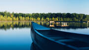 Une proue de canoë en bois au premier plan, flottant sur les eaux calmes du Lac de Grand-Lieu en Loire-Atlantique, avec une forêt dense se reflétant à l'horizon sous un ciel clair.